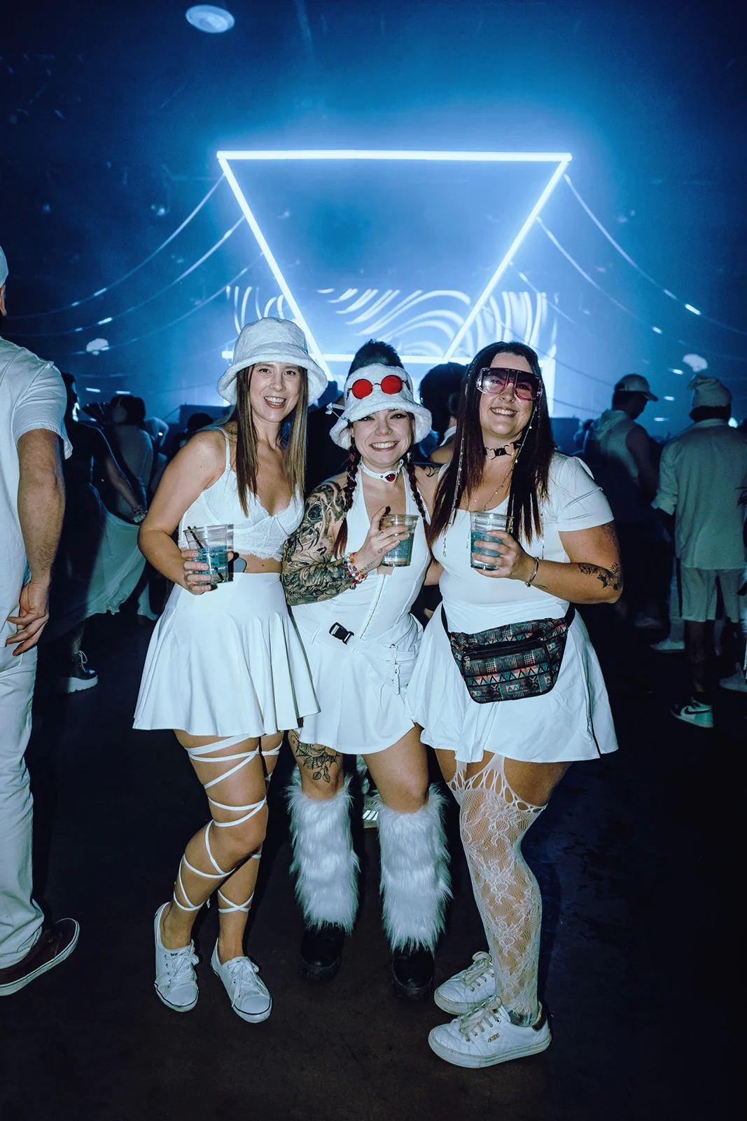 Three guests in white posing under a glowing blue neon triangle
