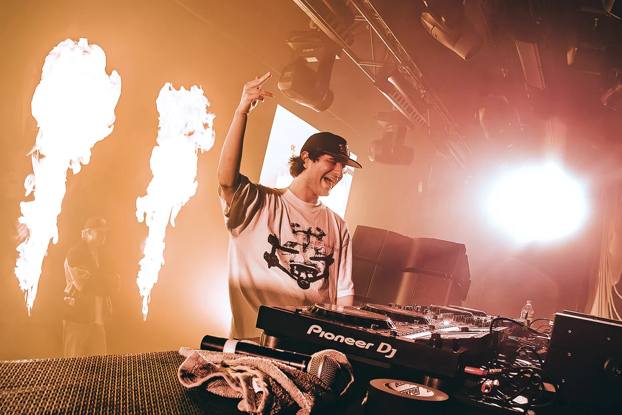A DJ raising an arm between columns of stage pyro